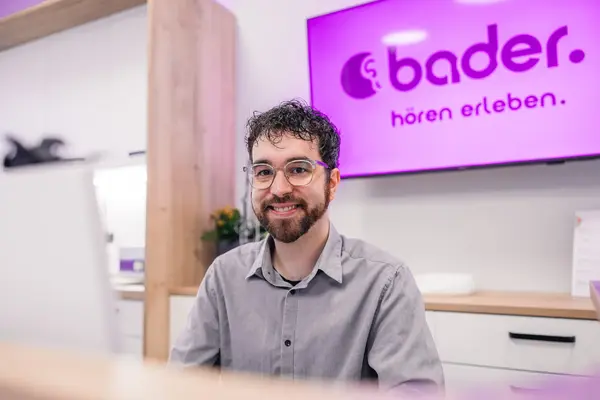 Lächelnder Mitarbeiter im Hörakustik-Shop vor bader-Logo und Beratungstheke.