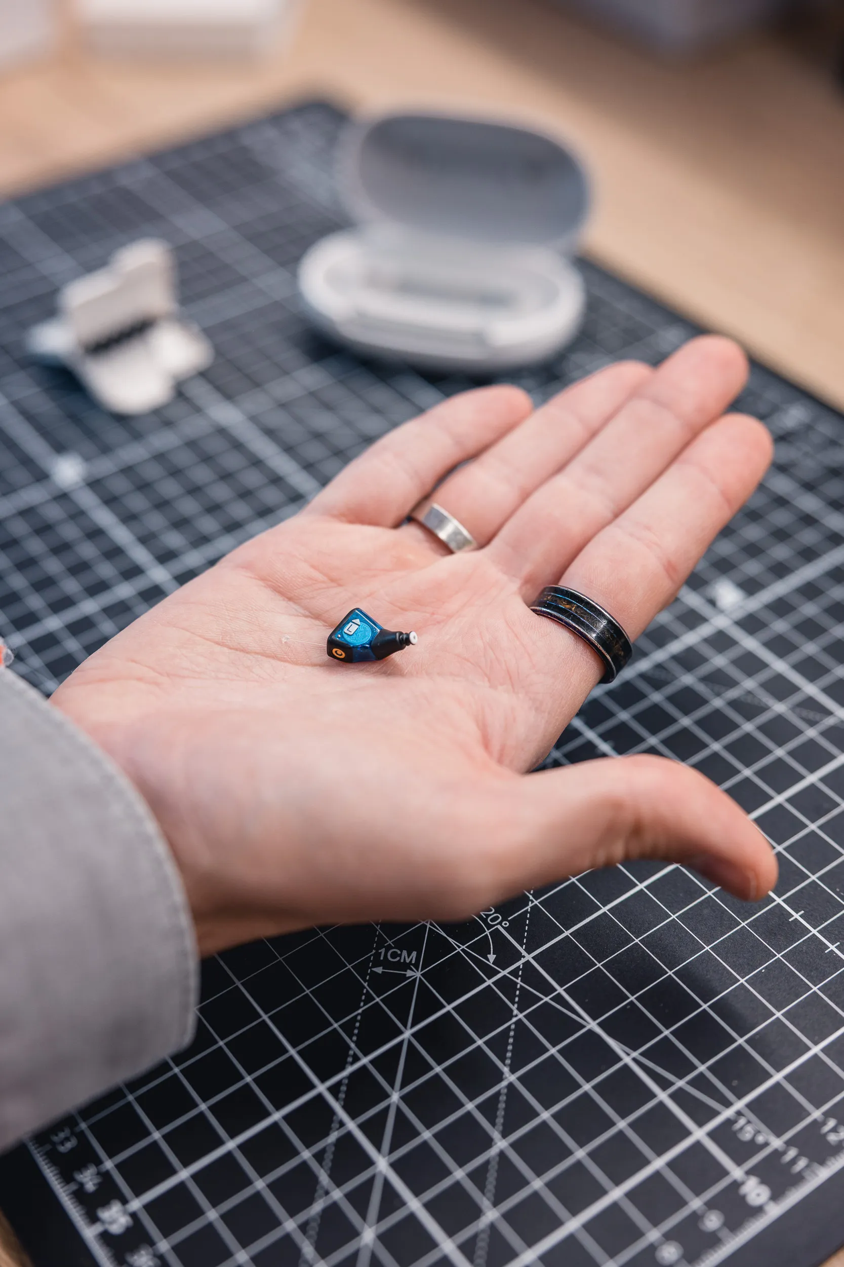 Hand hält kleines In-Ear-Hörgerät, im Hintergrund Ladecase auf Schneidmatte